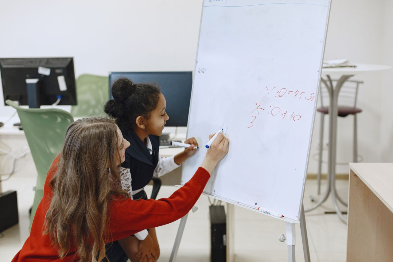 Home A teacher helps a student with math on a whiteboard in a classroom setting.